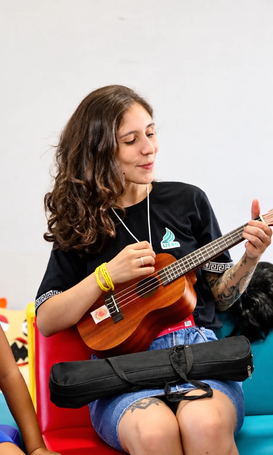 Karen toca ukulele sentada num sofá colorido, durante uma atividade com crianças.