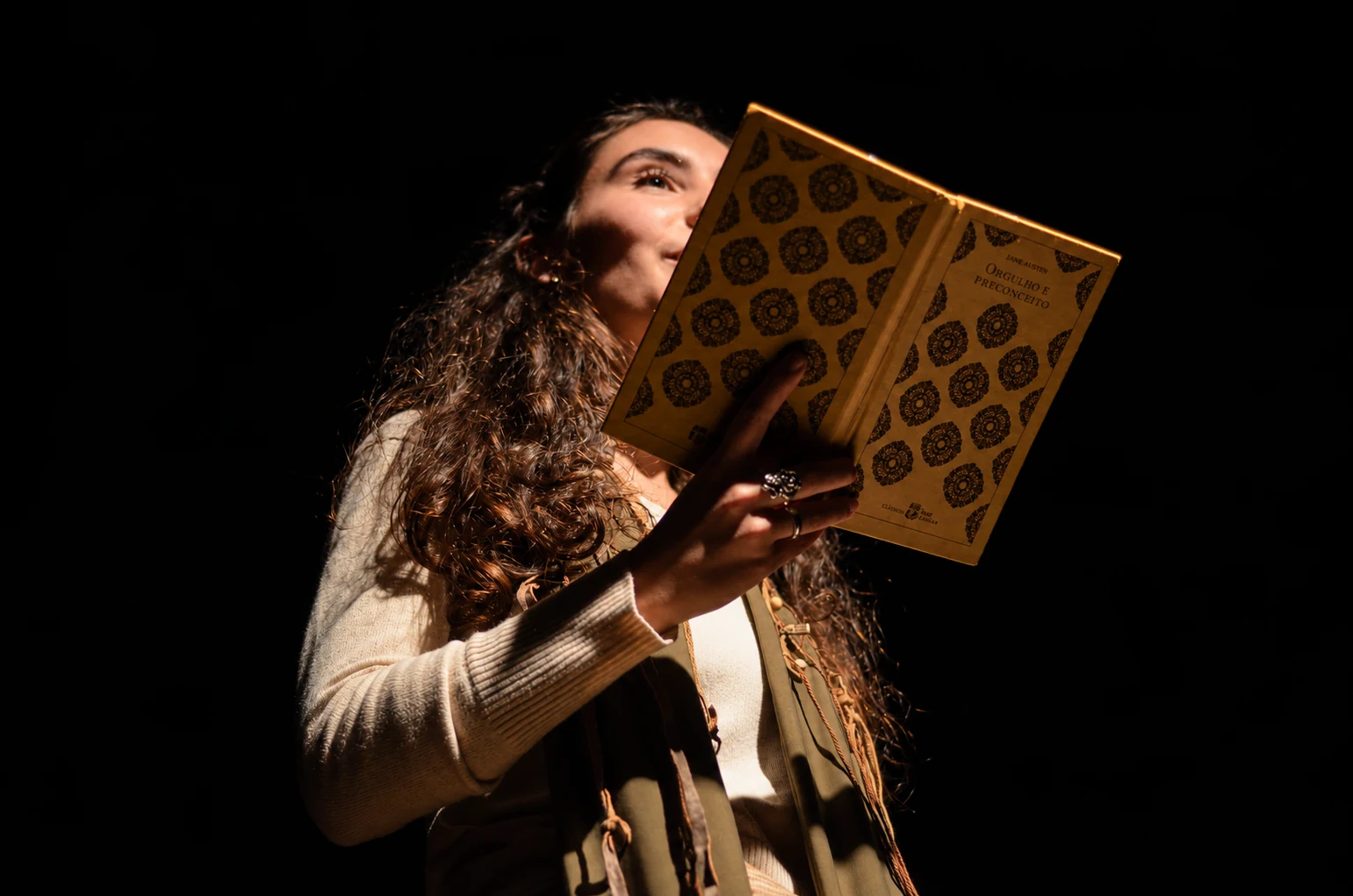 Karen em cena, sob um único foco de luz, lê um livro de capa dourada.