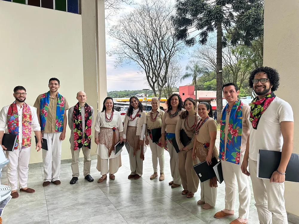 Grupo coral de roupas claras e lenços floridos, reunido num pátio coberto.