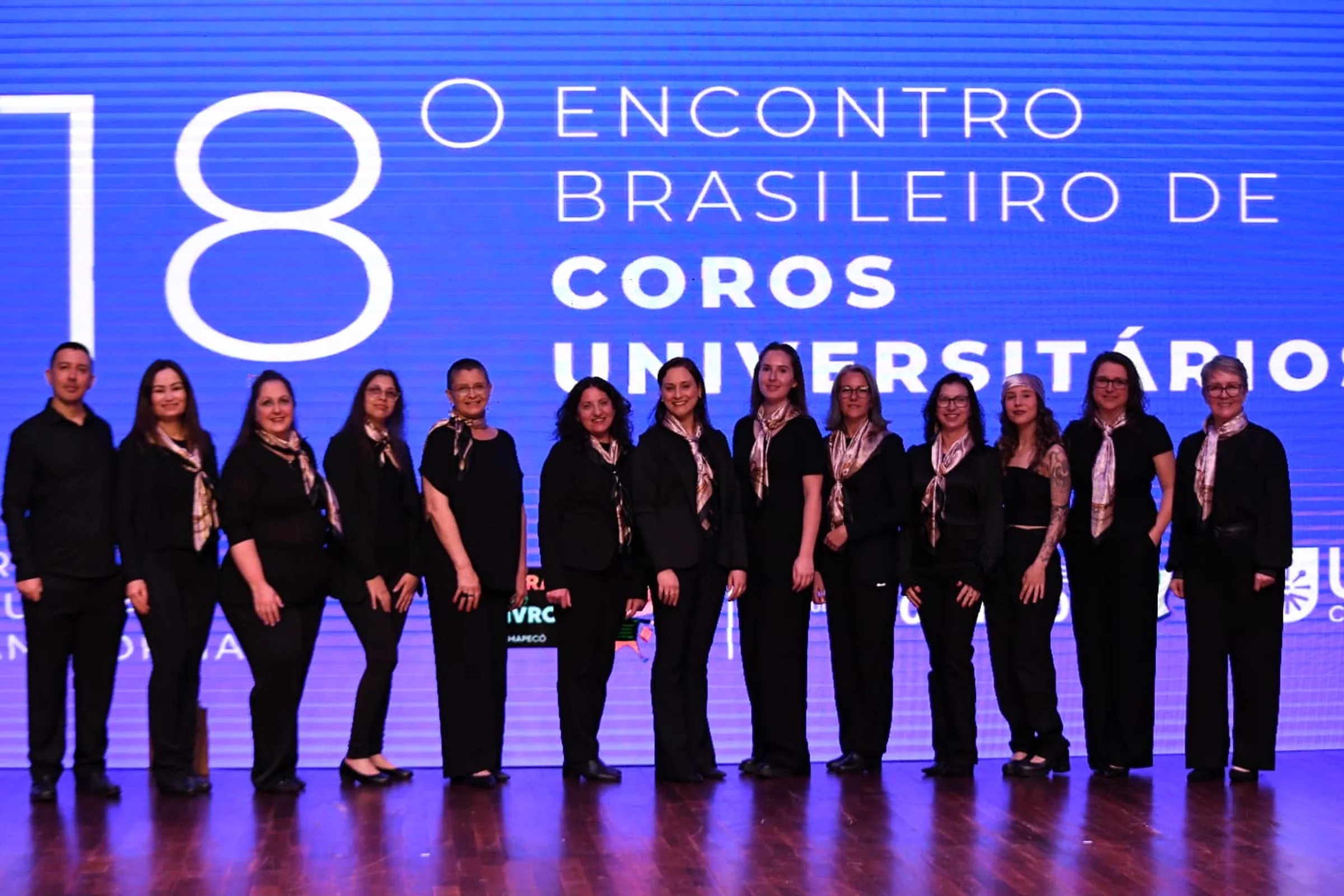 O coro completo em fila, de lenços estampados, diante do telão.