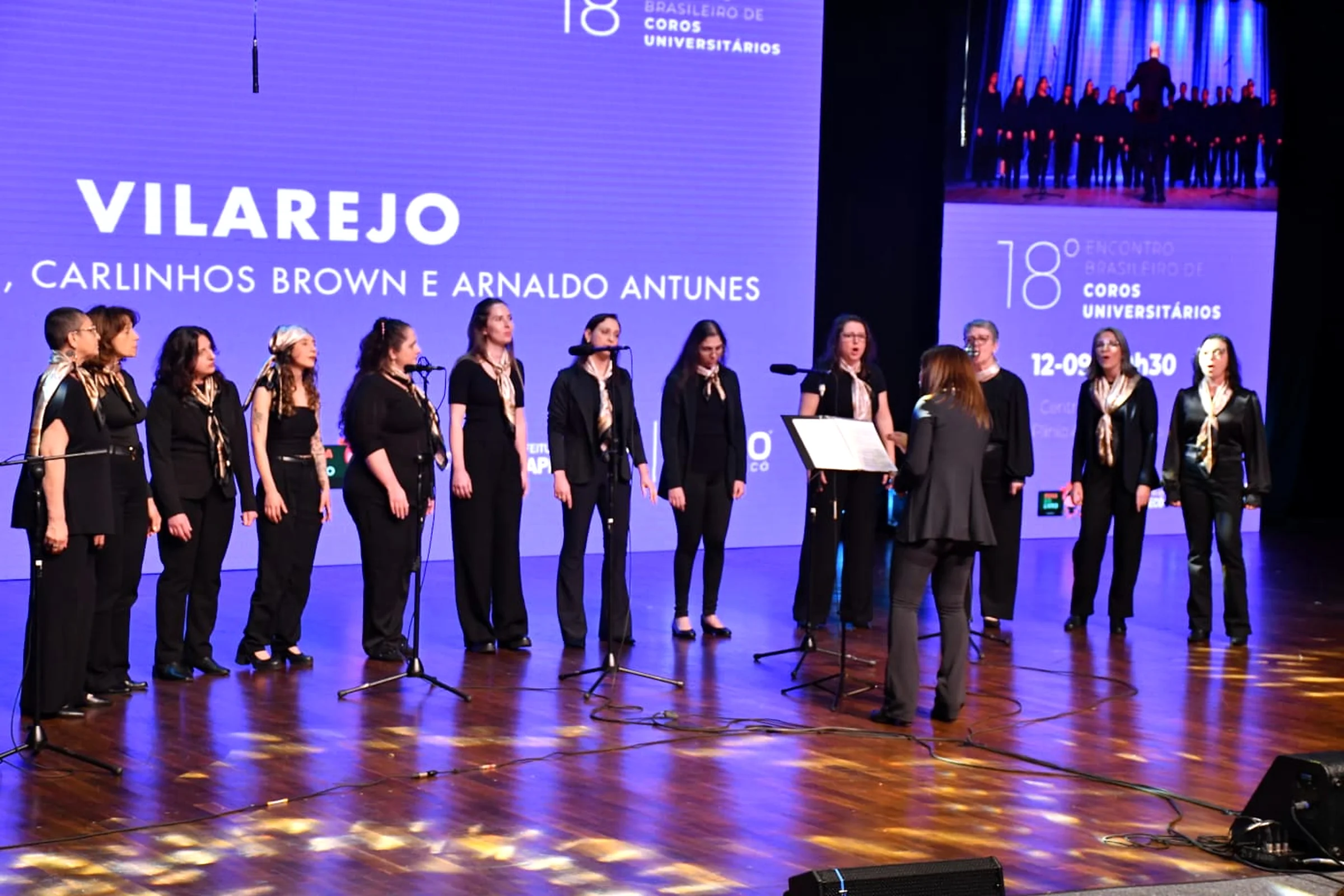 O coro no 18º Encontro Brasileiro de Coros Universitários, diante do telão com Vilarejo.